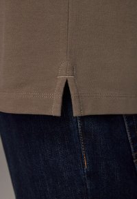 Ourlet de chemise en tissu brun texturé avec une petite fente latérale, portée sur un jean en denim bleu foncé avec des coutures visibles.