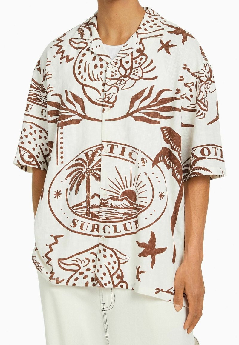 Camicia bianca a maniche corte con stampe tropicali marroni, con animali, piante e un sole. Design con colletto e bottoni sul davanti.