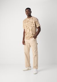 Beige gemustertes T-Shirt mit geometrischen Formen, kombiniert mit hellbeigen Hosen und weißen Sneakers mit beigen Akzenten.
