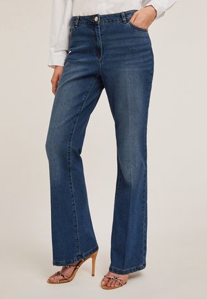 Vrouw draagt donkerblauwe jeans met rechte pijpen en voorzakken en beige hoge hakken sandalen, gecombineerd met een wit shirt met lange mouwen.