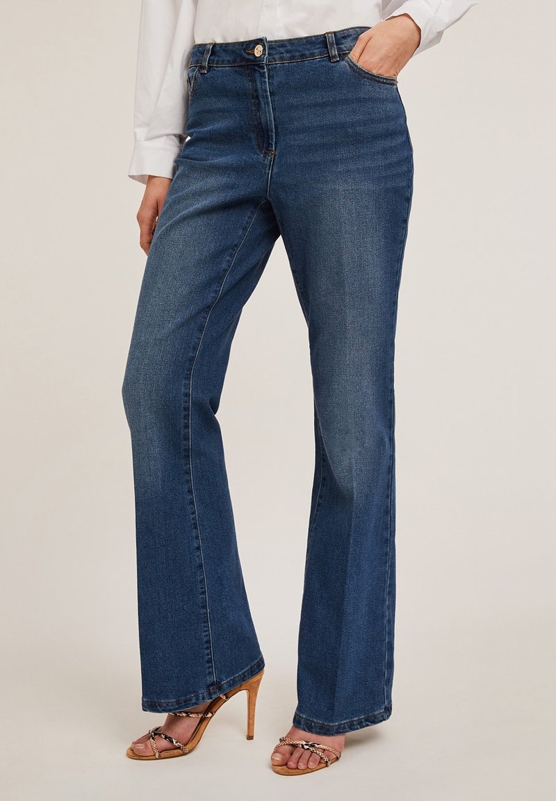 Femme portant un jean bleu foncé droit avec des poches avant et des sandales à talons hauts couleur camel, assorti à une chemise blanche à manches longues.