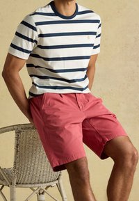 Homme portant un t-shirt blanc à rayures bleu marine et un short corail, assis sur une chaise tressée devant un fond beige.