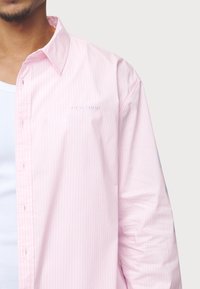 Jasnoróżowa, w paski koszula z guzikami o luźnym kroju. Posiada haftowane logo na lewej piersi oraz miękką, gładką tkaninę.
