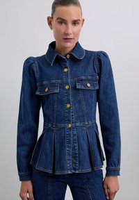 Denimjakke i mørkeblå med en fitted talje, peplumdetalje, guldknapaccenter og to forlommer. Lange ærmer med strukturerede skuldre.