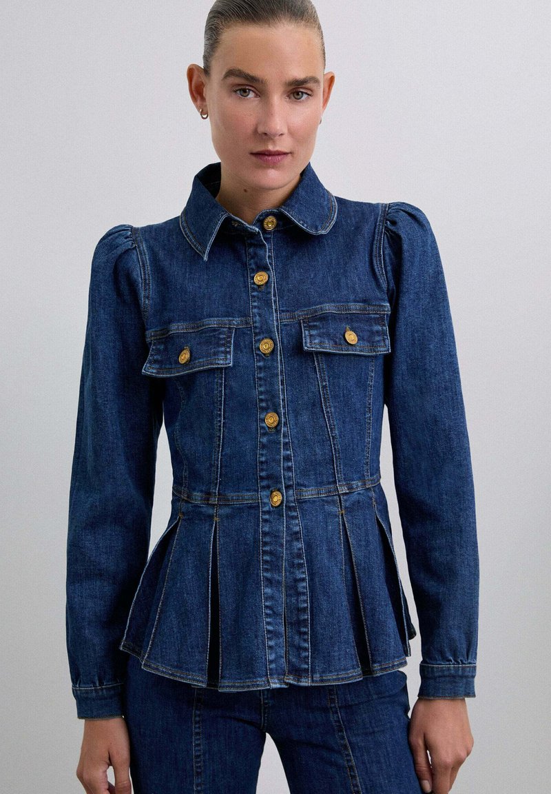 Denimjakke i mørkeblå med en fitted talje, peplumdetalje, guldknapaccenter og to forlommer. Lange ærmer med strukturerede skuldre.