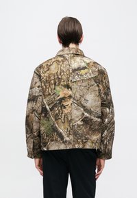 Μπουφάν καμουφλάζ σε γήινους τόνους με υφή που μοιάζει με φλοιό δέντρου και φύλλα, δύο πίσω τσέπες και κλασικό γιακά.