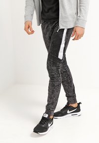 Černé jogger kalhoty s texturovaným povrchem, opatřené bílým bočním pruhem a panelovými detaily. V kombinaci s černými teniskami Nike.