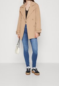Beige dubbele-breasted trenchcoat met brede kraag en knoopdetails, gecombineerd met blauwe skinny jeans en zwarte sneakers met plateausole.