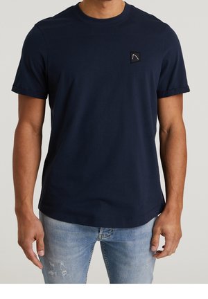 Mand iført en mørkeblå T-shirt med korte ærmer med et lille sort trekantet logo på brystet og lyseblå slidte jeans.