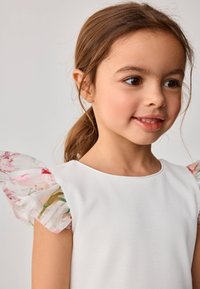 Fehér ruha áttetsző, virágmintás ujjal; puha textúra, kerek nyakkivágással; a háttér világos, semleges színű.