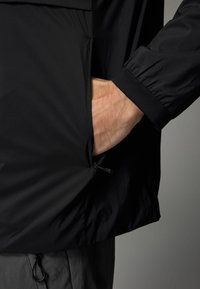 Juoda lengva striukė su užtrauktuku šone, elastingomis rankovėmis ir tekstūruotu audiniu. Apatinėje dalyje yra reguliuojama raištelių sistema.