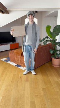 Egy személy szürke pulóverben, hozzáillő sapkában és sállal egy kartondobozt tart, amelyen az szerepel, hogy "Szeress. Viselj." egy fa padlós, kényelmes szobában.