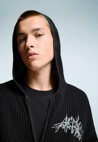 Fekete bordázott kötött kapucnis pulóver, cipzárral, amelyen fehér grafikai minta látható a mellrészen. Fekete kereknyakú póló felett viselve.