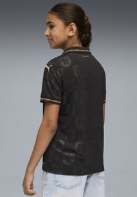 Črna polo majica z nežnim vzorcem ščita, s pisano črtasto ovratnico in zlatim logotipom na rami, izdelana iz lahkega blaga.