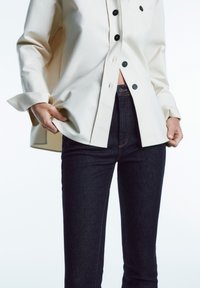 Kremowa koszula z guzikami w gładkim materiale z czarnymi guzikami, zestawiona z ciemnymi dżinsami z pomarańczowymi szwami i wysokim stanem.