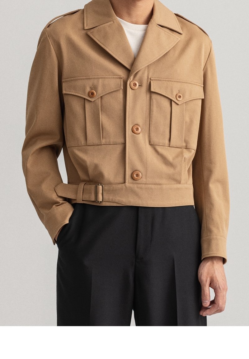 Homme portant une veste beige à boutons avec deux poches avant boutonnées, un détail ceinture à la taille et un pantalon noir.