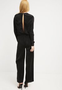 Mono negro con un acabado texturizado, mangas largas y un profundo escote en la espalda. Su diseño de pierna ancha fluye desde la cintura, acentuado por zapatos de tacón.