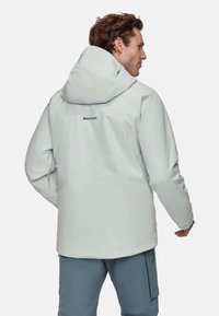 Veste imperméable bleu clair avec capuche, poches zippées et logo au dos. Texture lisse et design moderne ajusté.