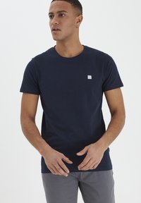 Jūras zils kokvilnas t-krekls ar apaļu kaklu un īsām piedurknēm. Uz kreisā krūškurvja ir neliels kvadrātveida logo. Parasts piegriezums.