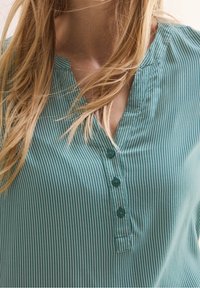 Teal stribet bluse med V-hals, korte ærmer og tre knapper foran. Har en blød tekstur og et tekstureret stofmønster.