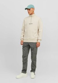Beige Reißverschluss-Sweatshirt mit schwarzem Logo, kombiniert mit grünen Cargo-Hosen und weißen Sneakern, ergänzt durch eine hellblaue Mütze.