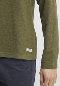 Olive green long-sleeved shirt with a textured fabric, featuring a small label on the hem and paired with dark pants.