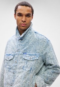 Man wearing a textured light blue button-up jacket with chest pockets, standing against a plain white background.