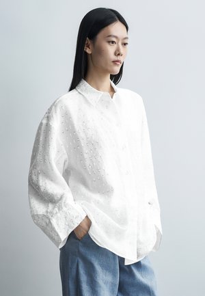 Jonge vrouw met lang zwart haar, die een oversized witte, gestructureerde blouse met knopen draagt en losse blauwe broek, staand met handen in de zakken.