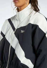 Mustvalge Reebok jakk kõrge kraega, lukuga sulgemine, diagonaalne värvi-blokeerimise disain, kerge ja elastne vöökoht.