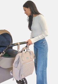 Bolso de pañales acolchado en color beige claro con dos asas, bolsillo frontal y cremalleras plateadas, sujetado a un cochecito al lado de una persona con jeans y una camiseta de manga larga.