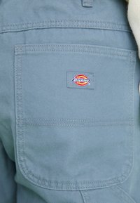 Blue fabric workwear featuring a large back pocket and a rectangular Dickies logo patch in red, yellow, and blue on the right side.