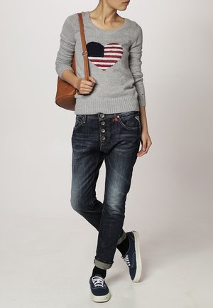 Osoba ubrana w szary sweter z nadrukiem serca, ciemne podwinięte dżinsy, granatowe trampki i nosząca brązową torbę na ramię.