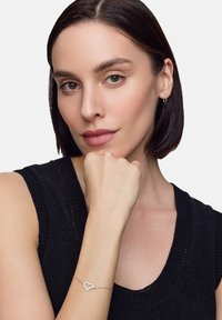 Srebrna bransoletka w kształcie serca z delikatnym łańcuszkiem, noszona na nadgarstku modelki, eksponująca gładkie, wypolerowane wykończenie na tle ciemnego swetra.