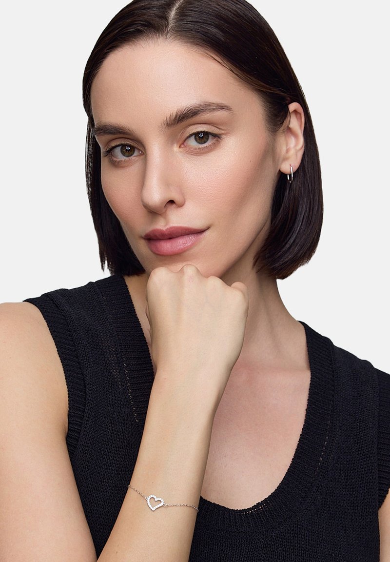 Srebrna bransoletka w kształcie serca z delikatnym łańcuszkiem, noszona na nadgarstku modelki, eksponująca gładkie, wypolerowane wykończenie na tle ciemnego swetra.