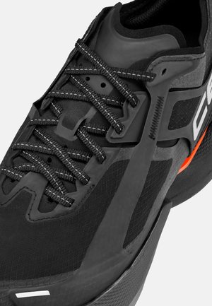 Close-up of a black athletic shoe with patterned black laces, textured fabric, and an orange accent near the sole.