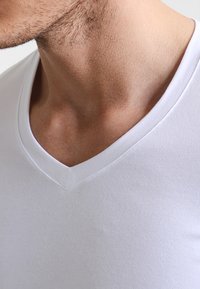 Biała bawełniana koszulka V-neck o gładkiej fakturze, z czystą linią dekoltu i minimalnymi detalami szwów, blisko szyi i okolicy ramion.