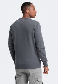 Sweatshirt gris à col rond avec des poignets et un ourlet côtelés, texture douce, et un détail de logo discret près du bas à gauche.