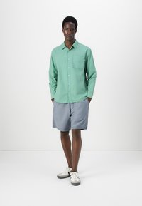 Camisa verde claro de botones con un bolsillo en el pecho, combinada con pantalones cortos grises y zapatillas. Tejido suave con un corte relajado.