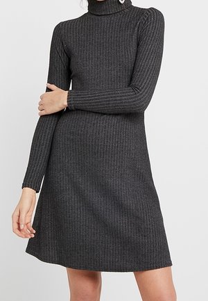 Ciemnoszara prążkowana sukienka dzianinowa z długim rękawem i golfem, noszona przez osobę z jedną ręką skrzyżowaną na klatce piersiowej.