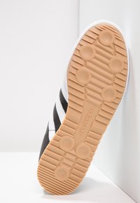 Černé tenisky s bílými pruhy, gumová podrážka s kruhovými úchyty a texturovaným povrchem. Logo je vyraženo na podrážce.