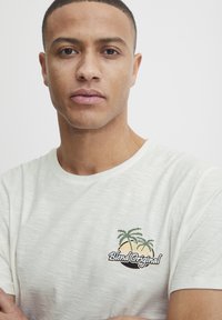 Hvid T-shirt med korte ærmer i en tekstureret finish, med et broderet palmetræ design og teksten "Blend Original" i sort.