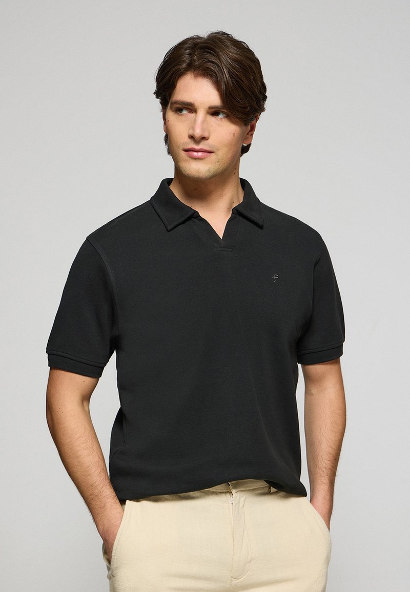 Svart polo skjorta med korta ärmar, mjuk textur, öppen krage, med en liten logotyp på bröstet. Kombinerad med ljusa byxor.