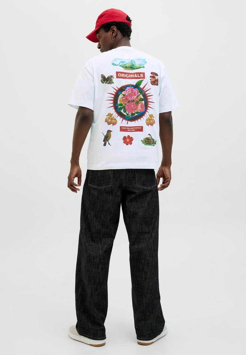 Camiseta blanca con un diseño gráfico colorido que presenta flores, animales y montañas en la parte posterior. Combinada con pantalones de mezclilla oscuros y zapatillas.