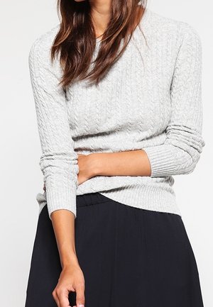 Kobieta ubrana w jasnoszary sweter w warkocze i czarną spódnicę, z jedną ręką skrzyżowaną na torsie.