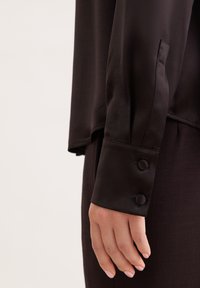 Primo piano di una mano che indossa una blusa a maniche lunghe marrone scuro con due bottoni rivestiti di tessuto sul polsino e pantaloni scuri abbinati.