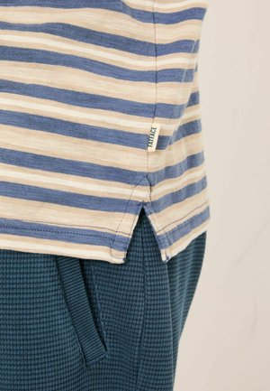 Stribet bomuld T-shirt med blå og creme farvede vandrette striber, med sideslidser ved kanten og et lille mærke med mærket. Båret over teksturerede mørkegrønne bukser.