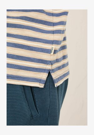 Gestreiftes Baumwoll-T-Shirt mit blauen und cremefarbenen horizontalen Streifen, ausgestattet mit seitlichen Schlitzen am Saum und einem kleinen Markenetikett. Trägt sich über strukturierten dunkelgrünen Hosen.