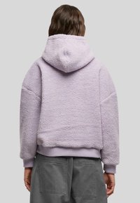 Lila cropped fleece hoodie met een gestructureerd oppervlak, grote capuchon en geribde manchetten. Gecombineerd met losse grijze broeken met zakken.