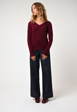 Maglione bordeaux a V, maniche lunghe, abbinato a jeans a gamba larga blu scuro. Modella che indossa scarpe con tacco maroon su uno sfondo neutro.