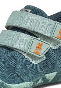 Scarpa in tessuto teal con motivo floreale testurizzato, dotata di due cinturini teal chiaro con testo in rilievo e un dettaglio con logo di coniglio arancione.